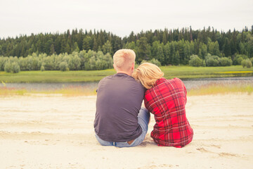A couple, a man and a woman, are sitting on the sand by the river. The concept of outdoor recreation, relaxation, love.