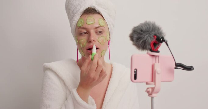 Women beauty blogging concept. Young female model does cosmetic makeup tutorial vlog poses at smartphone webcam applies hygienic lipstick cucumber mask wears bathrobe towel on head after taking shower