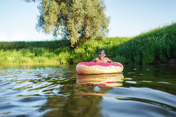 Happy boy in swimming glasses swims on an inflatable circle in the river in summer at sunset. A...