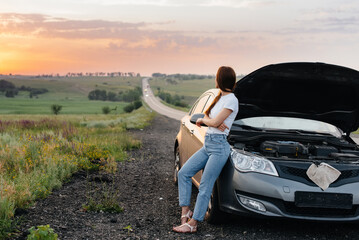 A frustrated young girl stands near a broken-down car in the middle of the highway during sunset. Breakdown and repair of the car. Waiting for help.