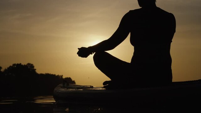 Silhouette Of Woman Relaxing In Lotus Pose On SUP Board On River At Sunset. Partial View Female Boarder Meditating In Beautiful Landscape. Paddle Yoga. Concept Of Healthy Lifestyle