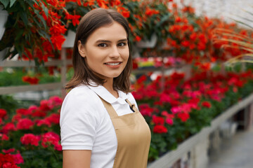 Close up portrait of smiling attractive young woman in special beige uniform standing in greenhouse on flowers background. Concept of work and care for flowerpots. 