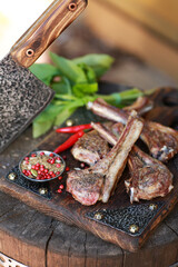 Grilled lamb ribs with spices and fresh vegetables and herbs on a wooden stump with a meat cleaver. Picnic, outdoor, sunlight, a square of lamb. Rustic. Background image, copy space
