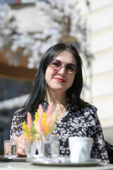 Smiling stylish woman in black shirt with sunglasses sitting at the table drinking coffee in the terrace of restaurant.