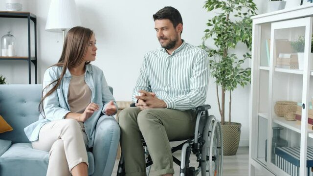 Disabled man in wheelchair is talking to woman smiling holding hands indoors at home. Physical disability and romantic relationship concept.