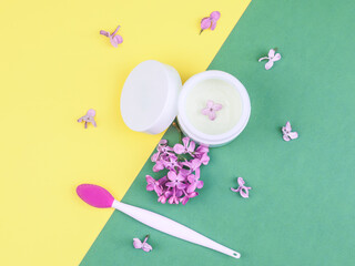 Face cream, lilac and mask brush.

Face cream, lilacs and a brush for masks lie on a yellow-green background, close-up top view.