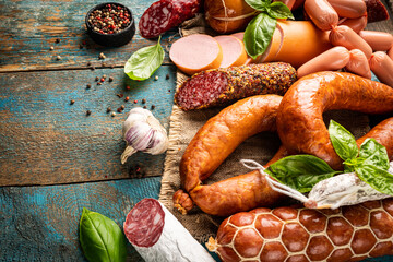 Set of different types of sausages, salami and smoked meat with basil and spices on wooden background. Top view.