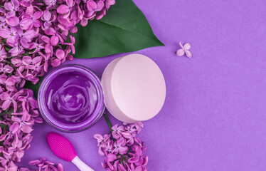 Lilac flowers and cosmetic cream.
Lilac flowers, cosmetic cream and a brush for masks lie on the left against a purple background with space for text on the right, top view close-up.