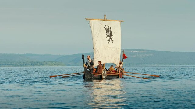 Vikings Sail on an Old Ship with a Raised Sail on a Calm River Against the Backdrop of a Rocky Coast. The Men Row the Oars Diligently Towards Adventure. Medieval Reconstruction.