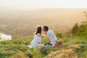 Beautiful young elegant couple enjoying picnic time on the sunset. They drinking wine and sitting on the hill.