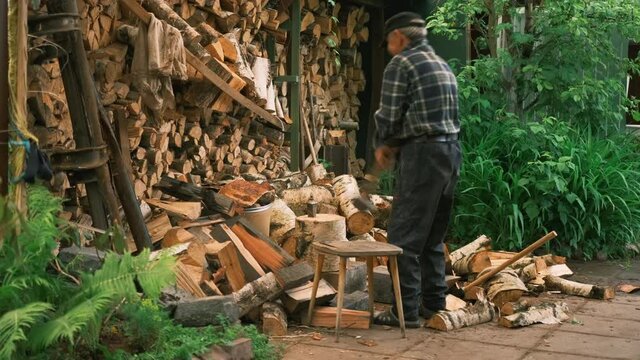 Side view an elderly man hits a wedge in a log with a sledgehammer and splits it. An elderly man in the yard next to the woodshed chopping wood in the summer. A man prepares firewood 