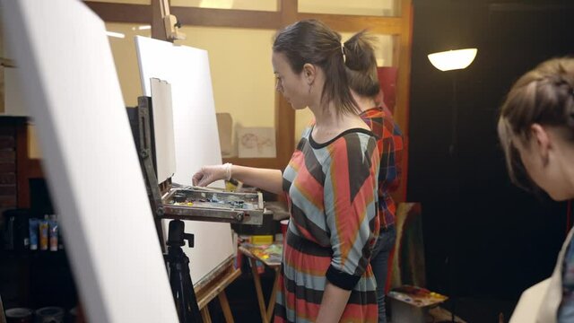 Women artists approaching the easel with paints and brushes to start painting.