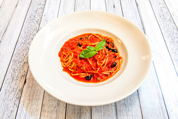 White plate with tomato puttanesca spaghetti and black olives with basil leaves on top