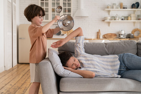 Naughty kid rumbling kitchenware while tired mother try to sleep on sofa in living room. Hyperactive preschool son make noises disturb young mom stressed and annoyed. Motherhood and children concept