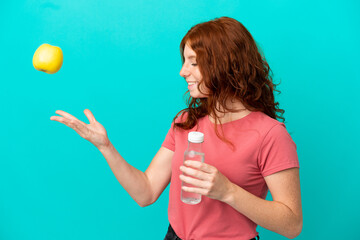 Teenager redhead girl isolated  on blue background with an apple and with a bottle of water