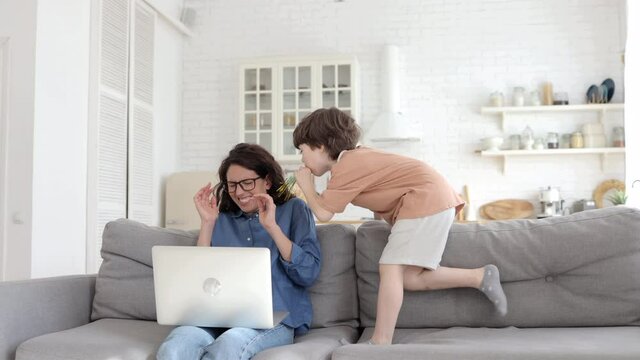 Bored hyperactive kid jumping and making noise at frustrated young mother trying to concentrate on freelance work in laptop on lockdown adn school quarantine. Mom parent tired of srubborn son at home
