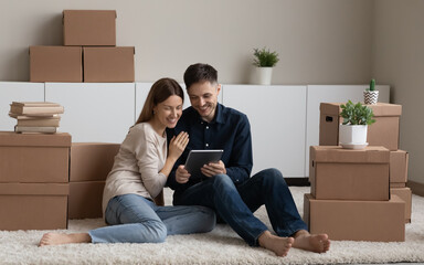 Smiling young Caucasian man and woman renters sit on floor in new home near boxed packages use tablet gadget. Happy couple tenants hold pad device browse internet search apartment for rent online.