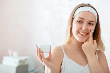 Young beautiful woman holds white jar of moisturizer cream applies cream on face in bathroom. Self...