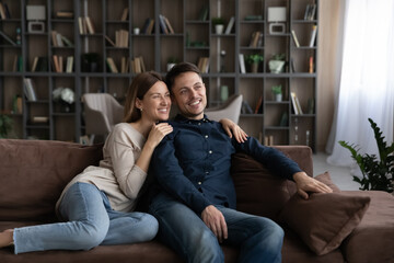 Smiling young Caucasian man and woman sit relax on sofa in living room look in distance dreaming planning. Happy couple renters rest on couch at home hug cuddle enjoy family life. Rent concept.