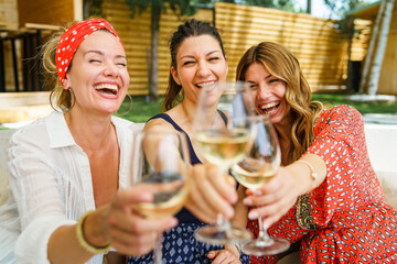 Three adult beautiful caucasian women female friends sitting outdoor in backyard with glasses of white wine having fun celebrating and drinking in summer day on vacation holiday - real people leisure - obrazy, fototapety, plakaty