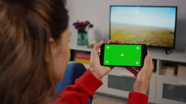 Phone Horizontal Green Screen For Copy Space Close Up Chroma Key Mockup. Back Over The Shoulder View Girl Watching Video Movie On Couch Home. Hand Phone Holding Technology Users.