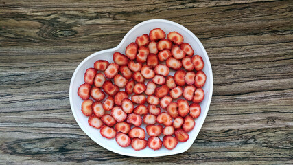 The sliced strawberries are in a heart-shaped plate. Romantic serving of a healthy dessert on a wooden background.