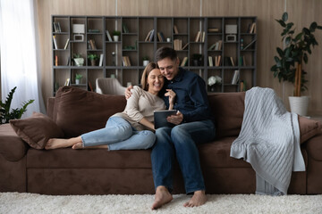 Obraz premium Smiling young Caucasian couple relax on couch at home talk speak on video webcam call on tablet together. Happy millennial man and woman rest on sofa in living room use pad. Technology concept.