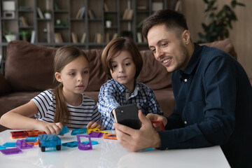 Fototapeta premium Happy young Caucasian father play toys with two teen kids use modern smartphone have webcam call on device. Smiling loving dad and children involved in funny game with blocks at home together.