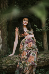 Pregnant likable Caucasian woman 25-30 year old wearing colorful dress and black hat posing outdoors in beautiful forest. Motherhood. Maternity. Autumn season.