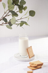 A glass of milk on a marble plate on a white table cloth, freshly baked cookies, branch of green eucalyptus