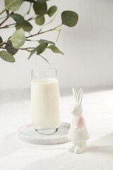 A glass of milk on a marble plate on a white table cloth, freshly baked cookies, branch of green eucalyptus , and white figurine of an easter bunny