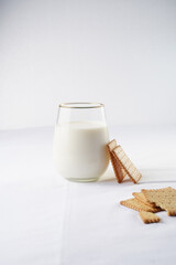 A glass of milk on the white table cloth and freshly baked cookies leaning on the glass