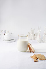 A glass of milk on a marble plate on a white table cloth, freshly baked cookies, and white easter bunnies, figurines