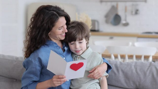 Caring son present mom greeting card for holiday event: mother day or birthday sit cuddling to mum reading postcard. Happy pleased young single mother hugging small boy on family holiday or valentines