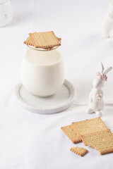 A glass of milk on a marble plate on a white table cloth, freshly baked cookies, and white easter bunnies, figurines