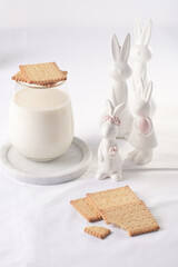 A glass of milk on a marble plate on a white table cloth, freshly baked cookies, and white easter bunnies, figurines