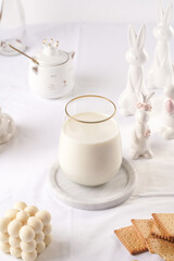 A glass of milk on a marble plate on a white table cloth, freshly baked cookies, creamy bubble candle and white easter bunnies, figurines