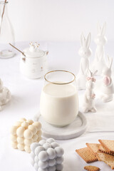 A glass of milk on a marble plate on a white table cloth, freshly baked cookies, creamy bubble candle and white easter bunnies, figurines