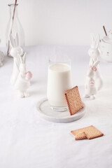 A glass of milk on a marble plate on a white table cloth, freshly baked cookies, and white easter bunnies, figurines