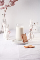 A glass of milk on a marble plate on a white table cloth, freshly baked cookies, and white easter bunnies, figurines