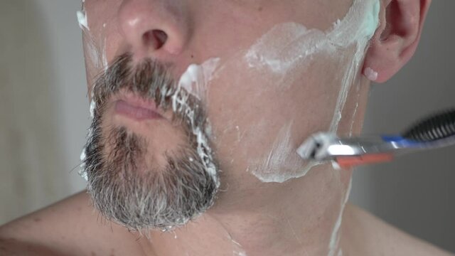 Middle aged man with foam on face shaving beard with a razor, close up 4K
