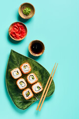 Top view traditional asian sushi rolls on ceramic decoration plate in shape of leaf with soy sauce, ginger, wasabi, chopsticks on blue turquoise background with copyspace.