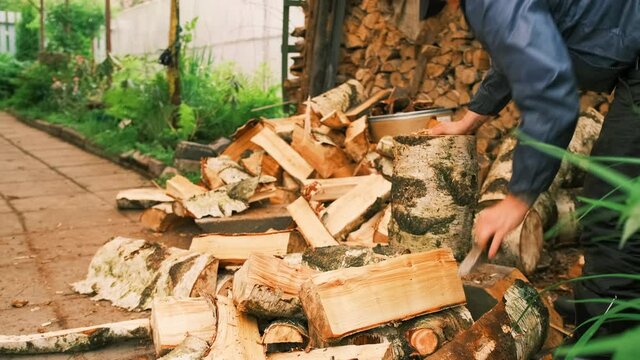 A young man is chopping wood in his courtyard of a country house. A man uses a hammer and a wedge to cut a piece of wood in half next to the firewood. Work in a country house. Village life