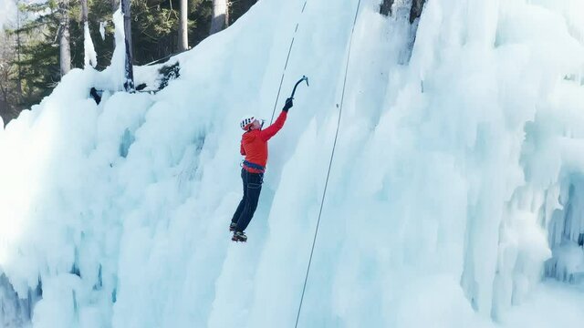Aerial shot of a Caucasian male ice climber in traction position, swinging ice axes overhead and planting the pick in the ice, side view
