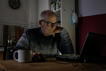 Old man with glasses works on his laptop in times of pandemic from the kitchen of his house, while having a coffee.