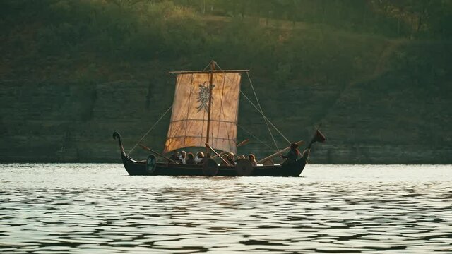 Vikings Sail on an Old Ship with a Lowered Sail on a Calmt River Against the Backdrop of Rocky Mountains. Concept on the theme of the Vikings and the early Middle Ages.