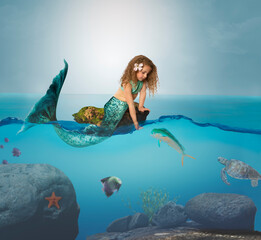 Curly hair little girl mermaid on floating rock in ocean feeding a fish - 442035953