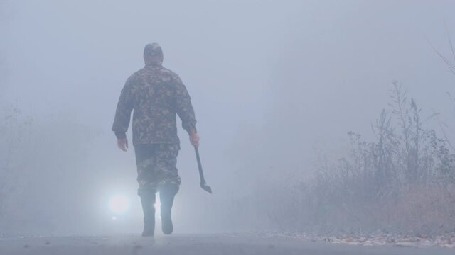 Aggressive man with an axe in his hand goes in the fog towards the car