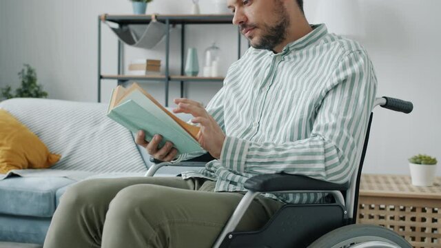 Portrait of handsome disabled man reading book sitting in wheelchair at home concentrated on interesting story. Disability and lifestyle concept.