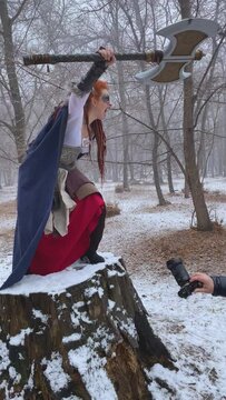 Male photographer shoots female Viking model in winter. Man in hooded jacket points camera at posing girl in historical clothing with robe and axe. Emotions on face. Large tree stump in snowy forest.